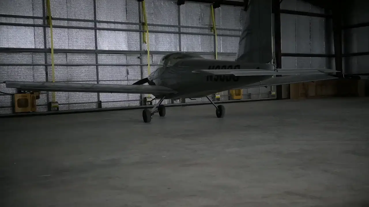 A low wing training airplane parked in a hangar as the hangar door rolls open behind it