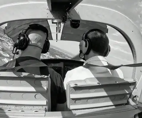 Black and white photo of two pilots in headsets seen from the back seat, flying over open country