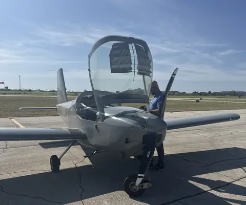 A pilot doing a preflight walkaround on a gray Van's RV-12 with the canopy open on a sunny ramp
